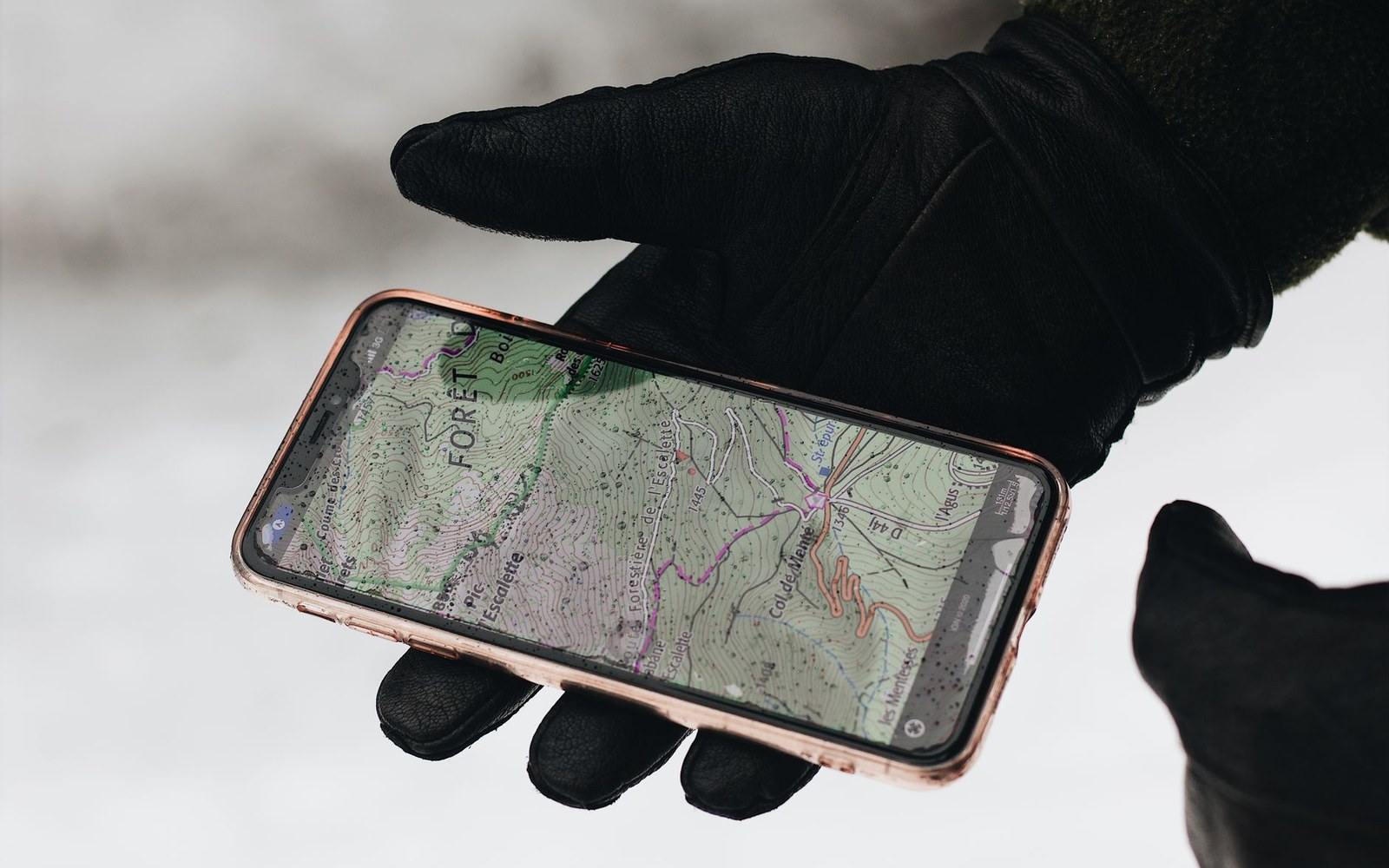 A gloved adult hand holding a phone displaying an avalanche forecast map, mountain terrain visible out of focus behind it