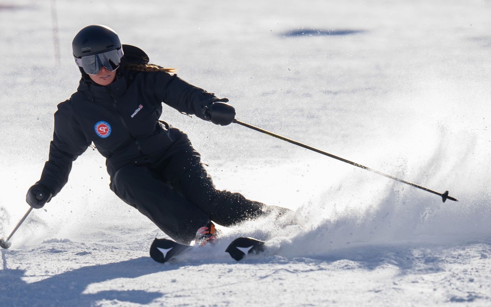 A skier in a black jacket and helmet carves through snow, spraying powder behind their skis