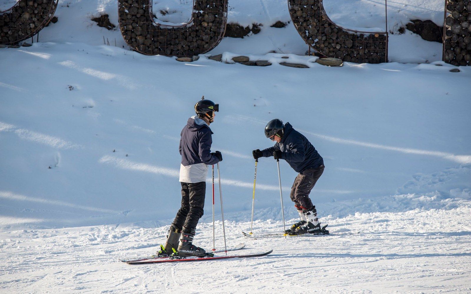 Two skiers with poles pause on a snowy slope beneath large stone-filled letter structures