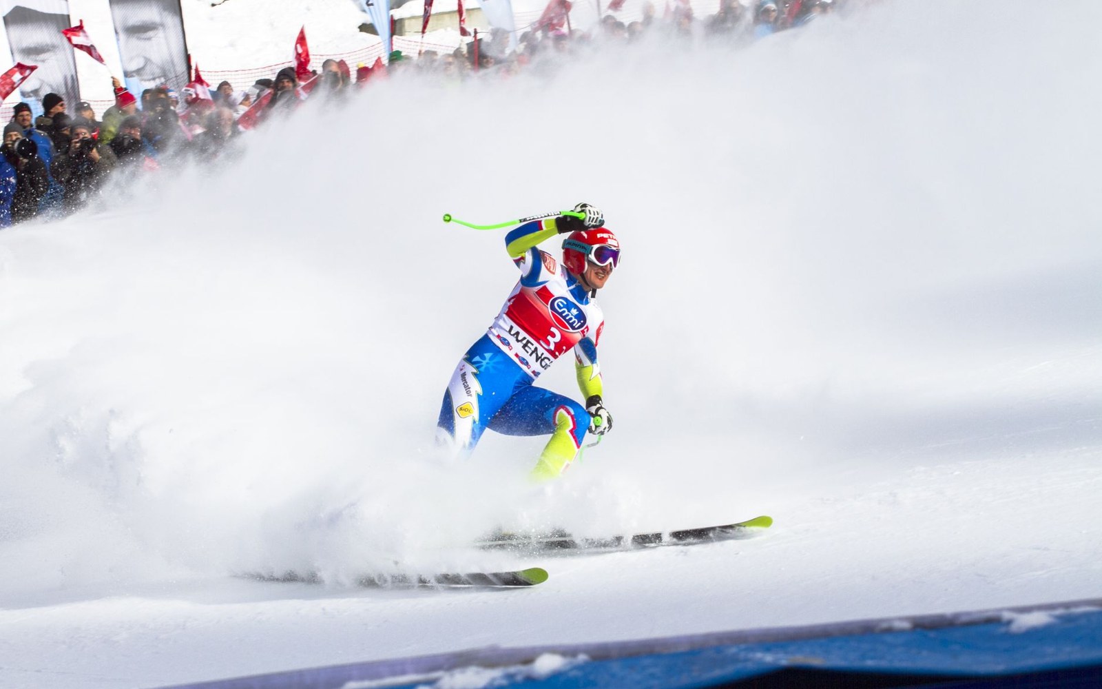 Skier throwing up a spray of snow while braking hard past finish-line crowds