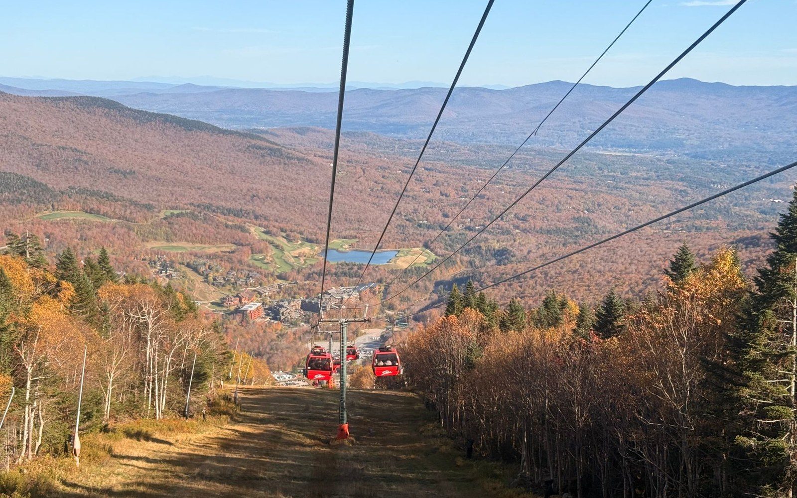 Red gondola cars descend a mountainside cable line above autumn forest and a distant lake