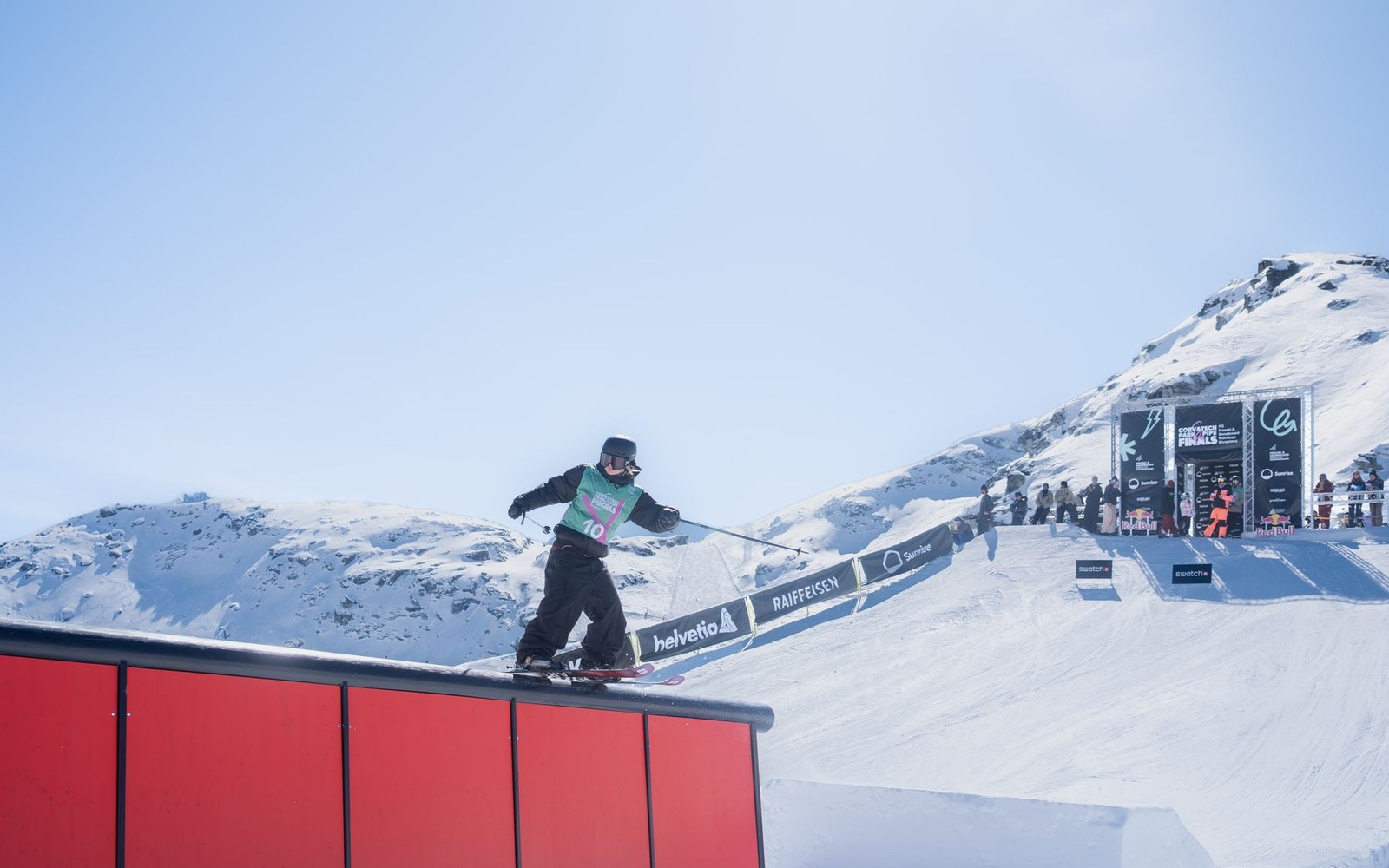 A skier slides along a red rail during a slopestyle competition with snowy peaks behind