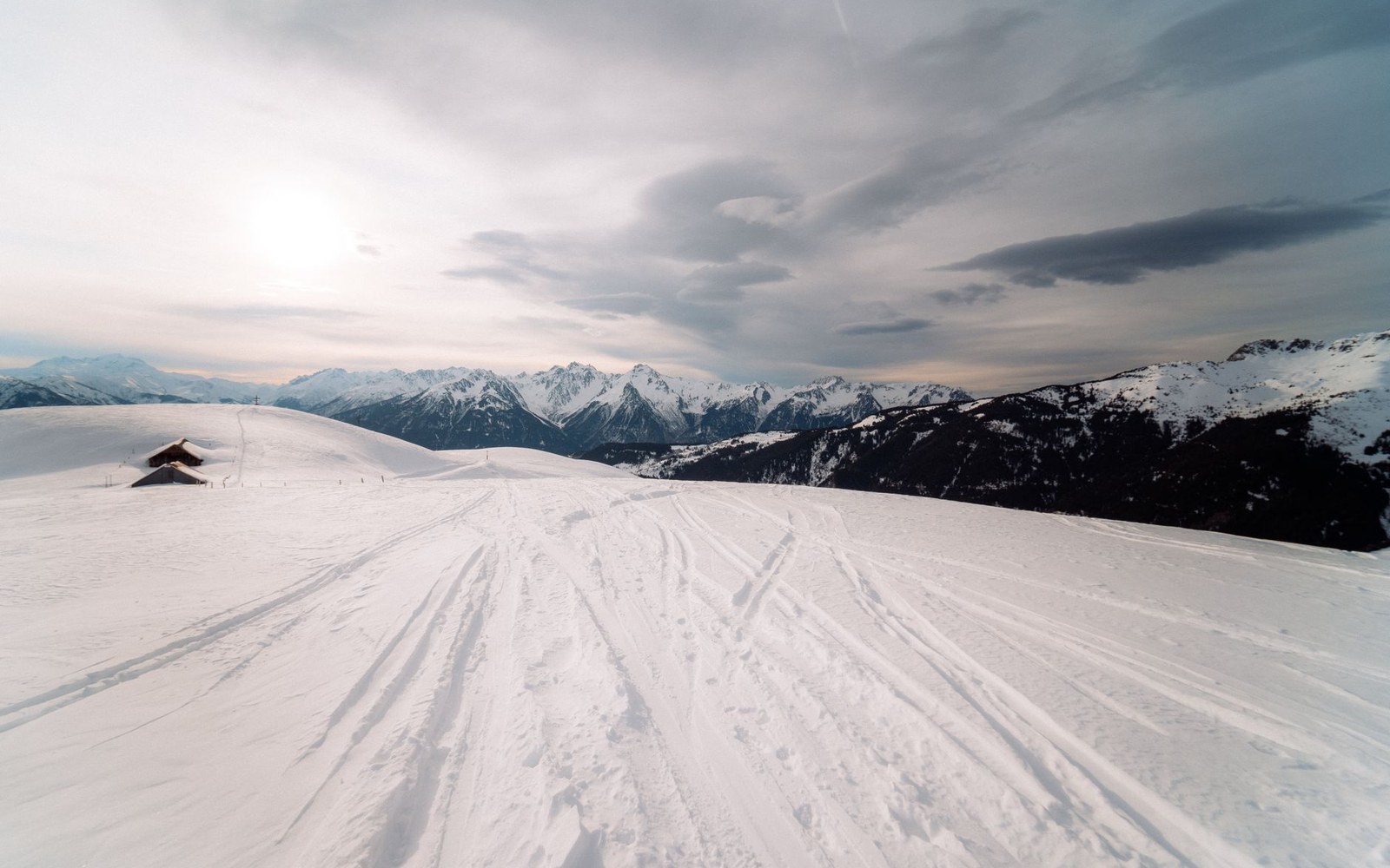 Snow-covered ski slope winds toward distant snowy mountain peaks under a cloudy sky