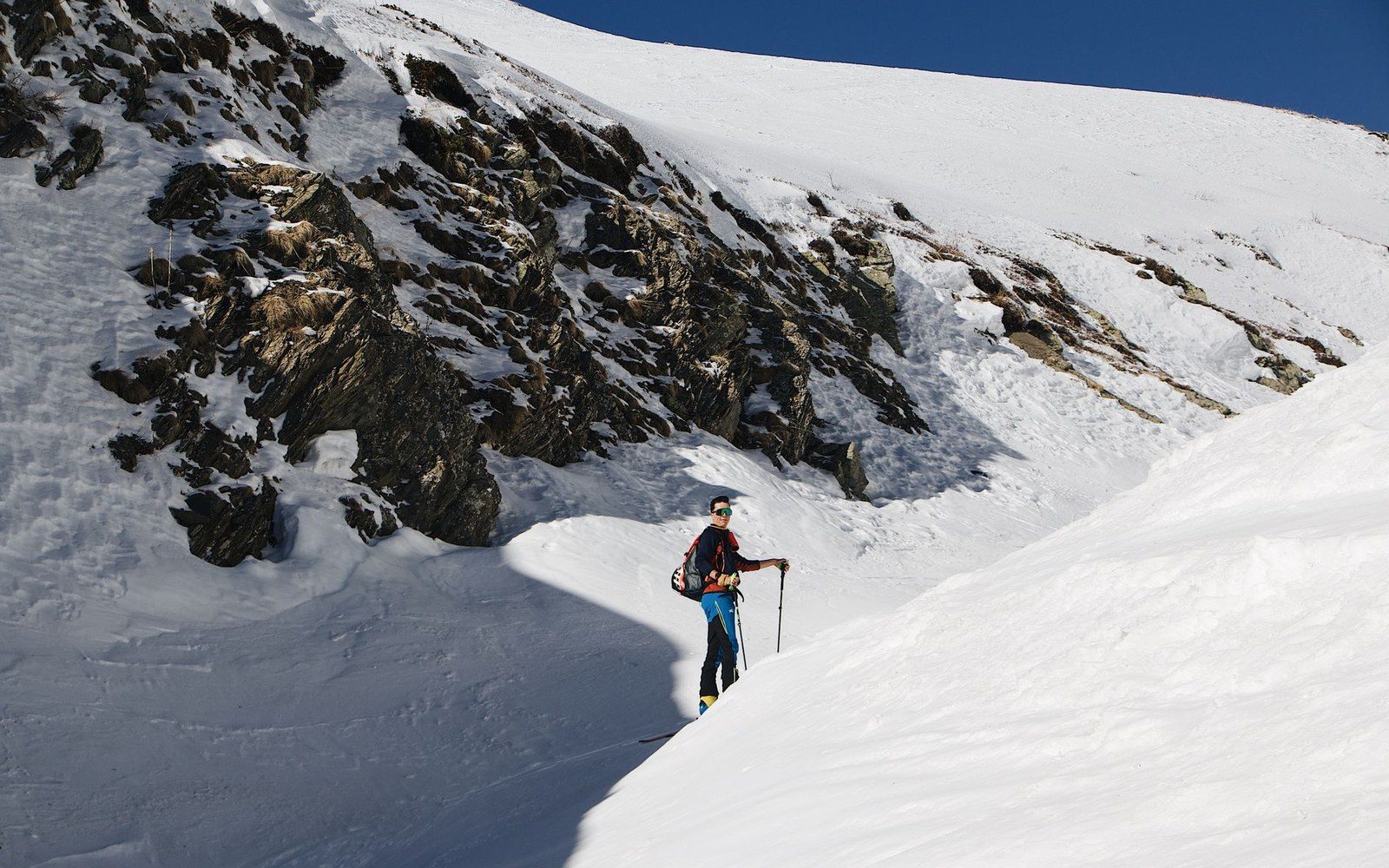 Skier with poles pauses on a snowy slope beneath rocky, snow-dusted cliffs