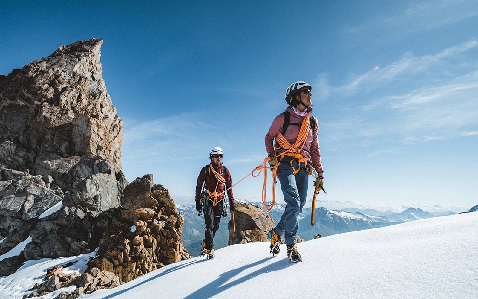 Two roped climbers in helmets and crampons walk along a snowy mountain ridge