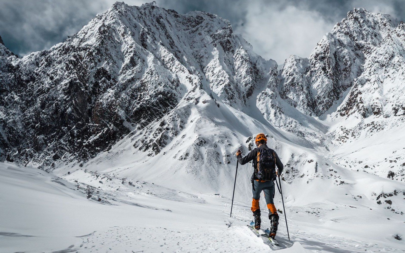Backcountry skier with poles and backpack facing jagged snow-covered mountain peaks