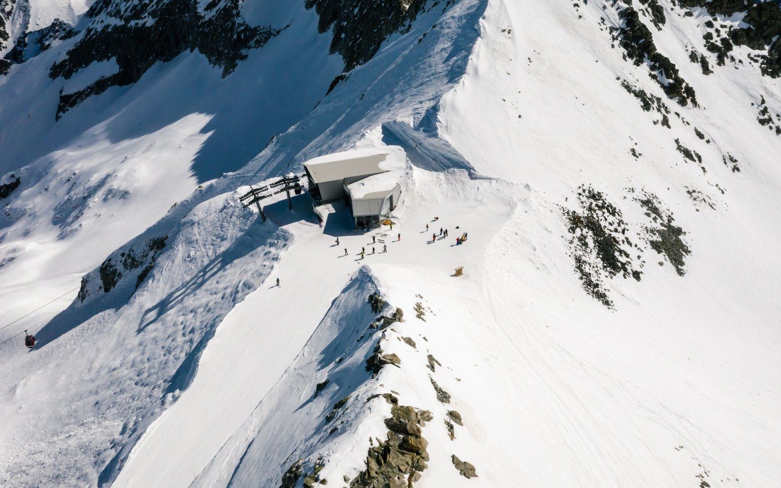 Skiers gather near a mountaintop cable car station on a snowy ridge