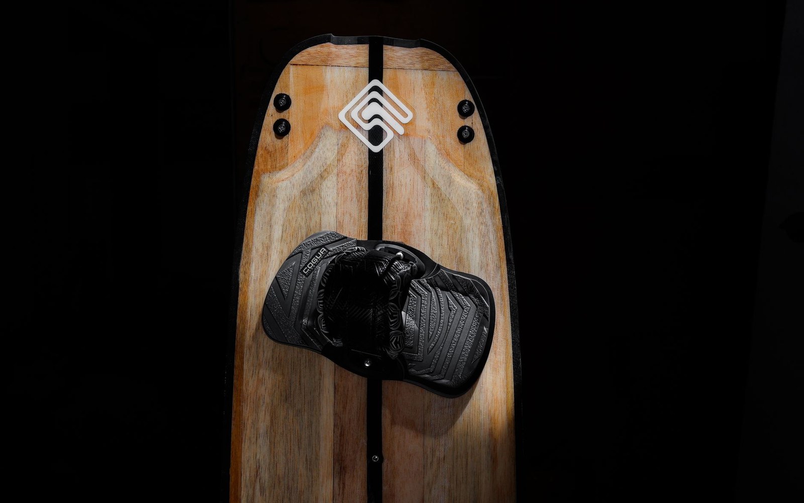A wooden wakeboard with a black boot binding and diamond-shaped logo, spotlighted against darkness