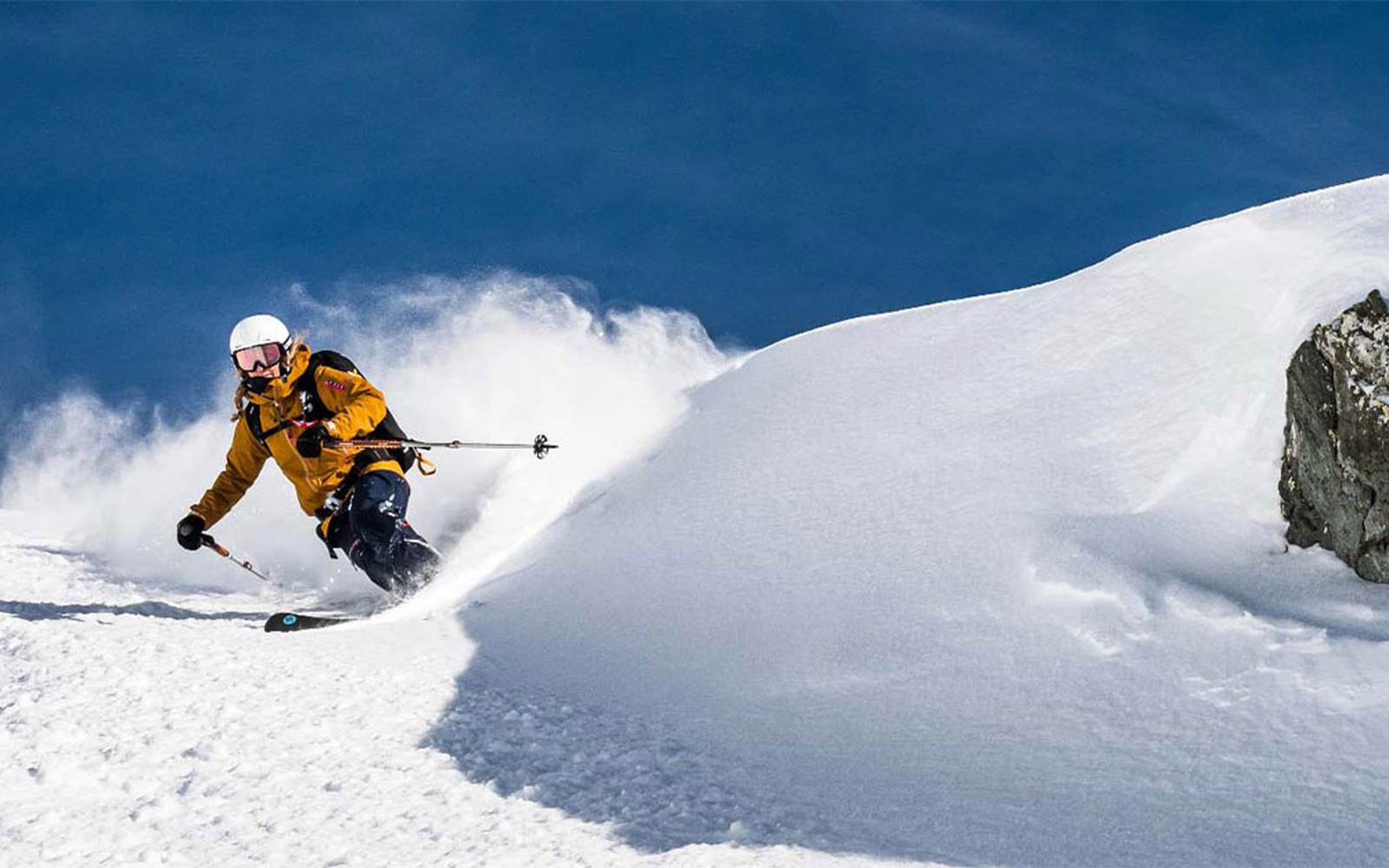 Skier in a yellow jacket carves through deep powder snow below a rocky ridge