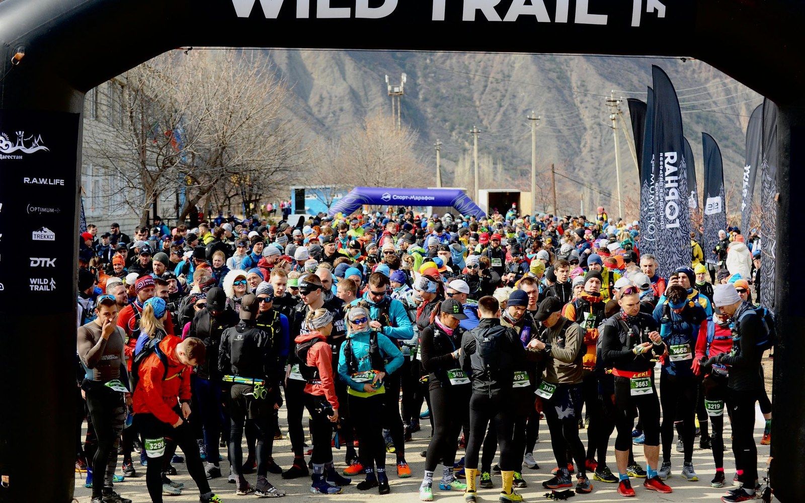 Runners with race bibs gather beneath a Wild Trail start arch before a mountain trail race
