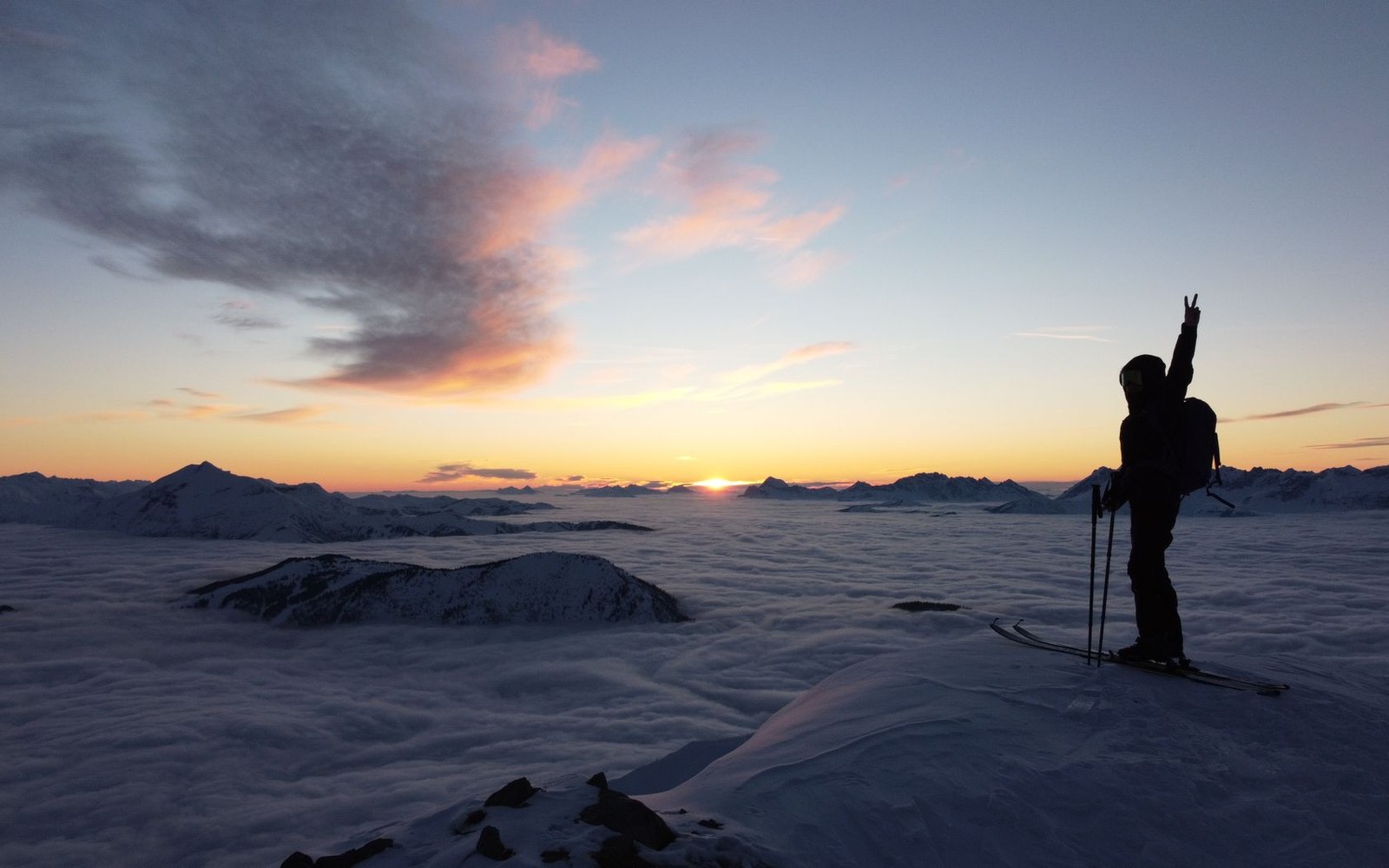 Skier raises a peace sign atop a peak above a sea of clouds at sunset
