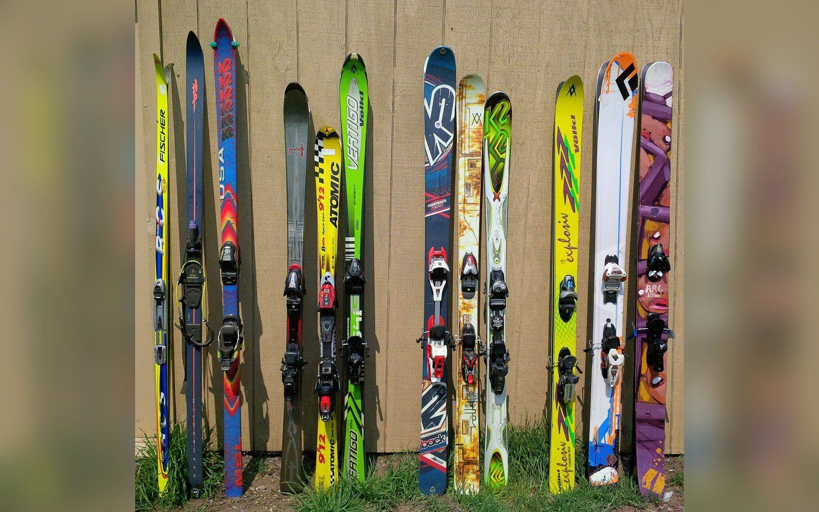 A row of colorful skis from various brands leaning against a wooden fence