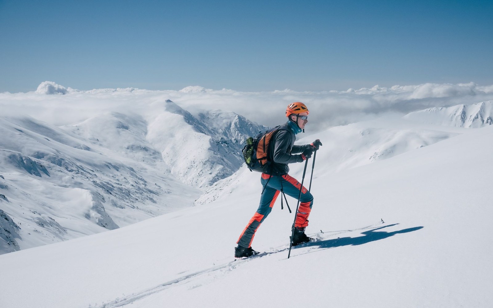 Adult hands peeling a climbing skin from a ski base at the tip, the skin's plush face visible, snow in the background